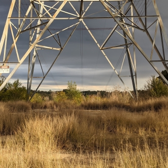 Transmission Tower View; sunlight on the grass and legs of tower in foreground and trees on either side in the midground, with dark clouds extending over the darkened valley in background