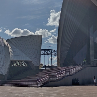 The Harbour Bridge Through The Opera House, Bennelong Point, with the flags atop the Harbour Bridge and five metal uprights visible between the Restaurant Shell on the left and the Concert Hall shell on the right, with flat walkway and low-incline steps below and brilliant blue sky above