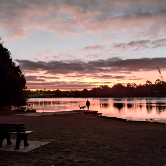 A Reflective Sunset, Yerrabi Pond, Gungahlin, with near-black clouds streaked left to right and bordered underneath by ochre in the sky reflected in the calm lake in the centre, with the silhouette of trees between the lake and the sky, with a tiny silhouette of a person and their dog standing front and centre on the shore of the lake, with grass stretching from the shore to the bottom of the frame