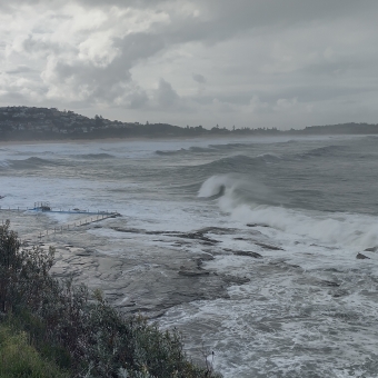 A Grey Outlook, South of Dee Why Beach, Dee Why, with dark grey water rolling into white foam beneath the cliff below and towards Dee Why Beach on the far left, with a rock pool barely visible behind a shrub covered cliff below to the left, with the horizon covered by a low headland chequered in houses and trees, with the clouds turning from silky clouds over the water on the right to sun-touched mashed potatoes over the land on the left