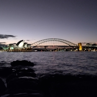 Mrs. Macquarie's Chair Lookout, Sydney, with a faded magenta-to-black sky in the background with a silhouetted outline of the Sydney Harbour Bridge at the centre and tiny skyscrapers of North Sydney on the right, with the Sydney Opera House lit up to the left, with a silhouetted tree close up on the far left of the image