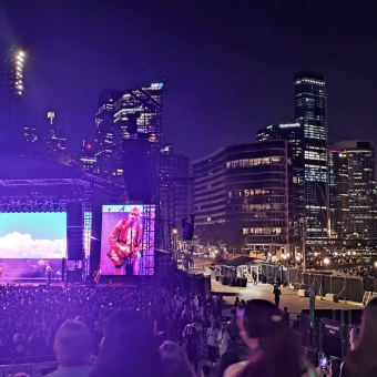 5 Seconds Of Summer concert, Sydney Opera House Forecourt, with the stage on the left of the image seeming to glow purple and blue with guitarist Michael Clifford projected on a big screen either side of the stage, with tall buildings behind Circular Quay outlined in black and chequered with many yellowed wndows, with a large crowd of heads streaming from the imeadiate foreground down to the stage who are covering all available floorspace