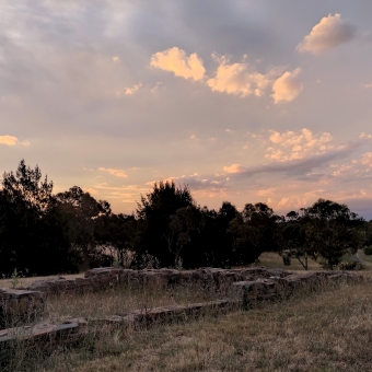 Crinigan's Stone Cottage, Amaroo, with a pink sunset dipping below the horizon which is hidden behind silhouetted fir trees, with the colour of the skies turning from pink to blue to almost white, with the stone remains of the cottage in the bottom left corner