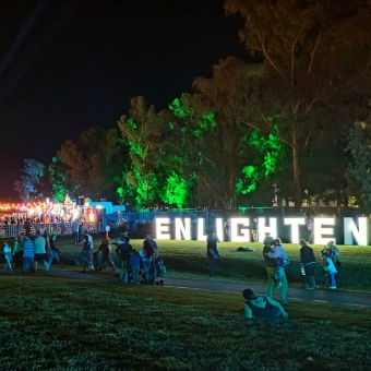 John Dunmore Lang Place, with on the right the word 'ENLIGHTEN' glowing in electrified letters the same height as people, with trees lit up with artificial green spotlights glowing through the leaves, with on the left red bar lights hung over the foodcourt and main stage, with powerful spotlights splayed into the sky behind it, with New Parliament House vicisble only by its flagpole visible in the distance