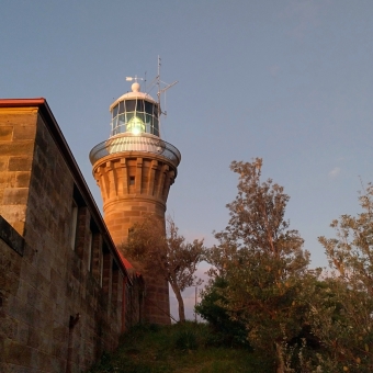 A Long Night Ahead, with the sandstone Lighthouse standing tall slightly to the left of centre, with the large light visibly lit on the inside, with a sandtsone wall on the left of the main lighthouse, with seemingly fluffy khaki-green trees on the right, with a clear blue sky in the background and the top half of the picture