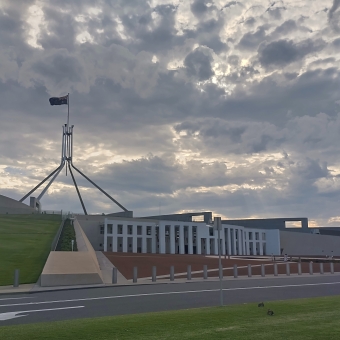 Parliament House, Canberra, with the spire and flag of Parliament House to the left of the frame and the front facing towards the right, with checkerboard clouds above gilded by sun and sun rays poking through at about the middle