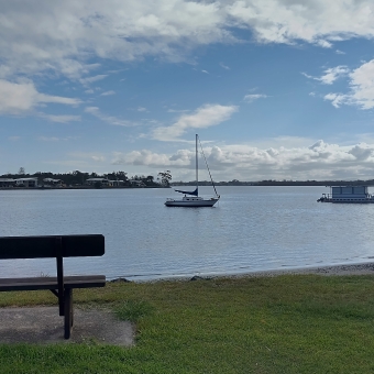 Stationed Yacht, Ferry Reserve, North Shore, with a small yacht in centre frame in the middle of a wide river, with a bench on the shore to the left of the image, with a floating dock in the water to the right of the image