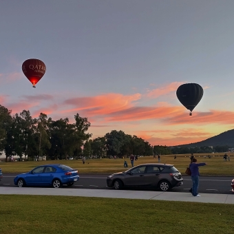 Canberra Balloon Spectacular, John Dunmore Lang Place, with a morning sunrise and peach-coloured streaky clouds along the horizon, with three hot-air balloons in descending order from left to right across the image