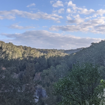 Hornsby Quarry; lower picture close small shrubs and grasses, extending to half way up to two low-rising hills covered in gum trees; top picture blue sky with clouds some fluffy some streaked across.