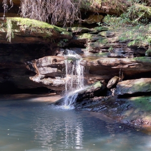 End-of-Track Waterfall 2: rocky cliffs stretch from top-left to bottom right with waterfall breaching at the centre falling onto the river banks and towards bottom left pooling into murky mix of dark greens and blues.