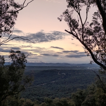 Sunset from the north side of Galambary, with dark green trees silhouetting left and right of the frmane which extend down and up the next ridge to the horizon, with peach and orange coloured sky streaked with grey clouds getting closer together the closer they are to the horizon