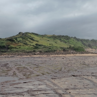 Long Reef Headland, Collaroy, with a moss-green hill stretching across the centre of the picture and into cliffs on the right, with an almost flat rock shelf carved with straight lines below, with silky grey clouds which have an eerie almost-aquamarine tinge above