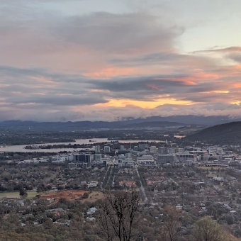 Canberra Under Fire, with Parliament House and ANZAC Parade on the far left and Caberra Central in the middle and Telstra Tower on the right, with the sky covered in streaky clouds mixing between glowing orange and dark magenta haze with the whole landscape especially Lake Burley Griffin a muted hue