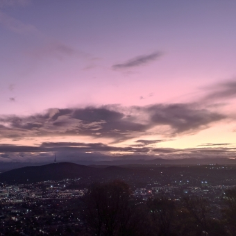 A Stratospheric Glow, with some patchy and streaky clouds silhouetted by a glow emanating from the hoizon which turns from orange through magenta through purple to blue the further from the setting sun, with the land below dotted with a multiude of tiny lights which are actually buildings