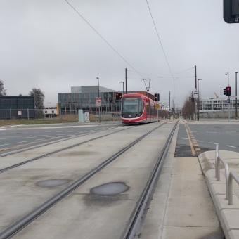 Gungahlin Centre Train Tracks, Gungahlin, with a red tram at the centre of the the image trailling away into the distance, with the dual tracks it's on coming into the fore, with low high-rise buildings left and right and a road crossing the mid-line of the picture