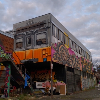 Train Graffiti, Watson Street, Turner, with a building taking up most of the photo which is covered in graffiti, with the most dominant piece being an overhanging side of the buildng made to look like a Sydney-based S-Set train which is also covered on its lower portion with graffiti tags, with the normal suburban street below disappearing off to the right and into the distance, with the mottled sky covered in pale blues and greys