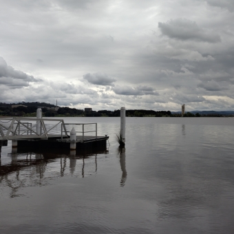 Solitary Wharf, Lake Burley Griffin, Canberra, with a walkbridge to a small pontoon in the centre of the water in the bottom half of the image, with the grey clouds looking like chequered mashed potato also reflected by colour in the water, with the divide between water and sky a silhouetted opposing coastling with the National Carillon just visible to the right of the picture