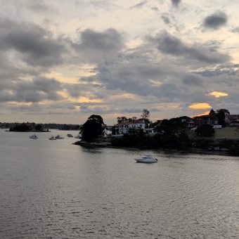 Celebrated Sydney Sunset, Aqua Luna Waterfront Dining, Drummoyne, with rippled water and chequered sky divided by thin coastline, with the setting sun glowing golden behind the patchy and grey clouds on the right side, with the grey and gold and faded blue reflected in the scaly water below, with the opposing coastlines dotted with many trees and a few buildings