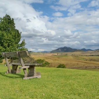 The View Over Gloucester, Kia Ora Lookout Retreat, Kia Ora, with a seat in the left sde foregrounds which is in front of a large and green tree, with the seat facing the right, with the centre and right fading into a valley and further mountain in the distance, with the valley dotted with faded grasses, trees, and houses