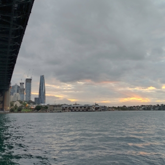 Milson's Point under the Sydney Harbour Bridge, with the under-side of the Harbour Bridge covering the sky on the left, with thick then thin mashed potato clouds over the sky which have enough gaps for a brilliant orange sunset stream through the clouds towards the Harbour Bridge, with the CBD buildings including Barangaroo visible betwixt the Harbour Bridge and the sunset, with the rippling water below reflecting grey with hints of teal