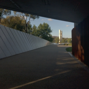 National Carillon, Lake Burley Griffin, Parkes, with the Carillon standing in the bright afternoon sun immediately surrounded by the bluest sky and greenest shrubs but further framed by a walking path and a retaining wall and an overpass which extend to the edges of the picture which all also become darker the closer to the edge they become,  with only a sliver of green trees visible above the retaining wall which seems to become a larger gap the closer it gets to the Carillon