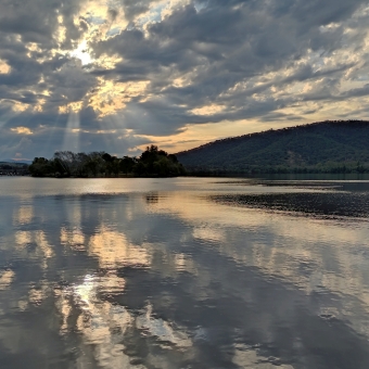 Reflected Sunrays, Lawson Crescent Viewing Deck, Acton, with orange sunlight streaming through fluffy and broken grey and gilded clouds, with the almost still water below reflecting the sky as though tiled, with tall hills in the far distance coming slightly closer to Black Mountain and Telstra Tower, with all these mountains just covering the horizon