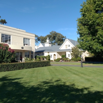 Other Galleries, with a meticulously mown lawn below, with a Horse Chestnut tree - also known as a Conker Tree - on the right, with a Gallery Building on the left which is protected by a low hedge and a red-flowering shrub, with a blue sky above, with another building reminiscent of a modern-traditional church in the centre