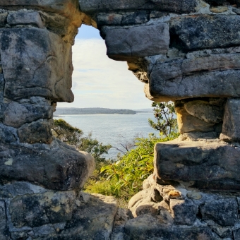 Coastal View, North Fort Wall, Manly, with the view an embrasure framed by grey weathered sandstone which is shaped like a pixellated kite, with Freshwater Beach and Freshwater Headland and the southern tip of South Curl Curl Beach visible in the disnace across blue ocean which is in part covered by green shrubs and ferns in the near-ish foreground, with thin grey clouds near the horizon only visible which covers an otherwise blue sky which is not visible in the picture but implied by the lighting