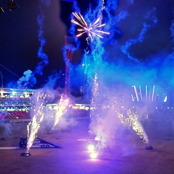 Fireworks Finale, with a darkened stadium lit up by fireworks and coloured spotlights, with the centre of the stadium containing seven fireworks shooters which have six in a large circle and in the centre, with the circle shooting out bright fireworks in a 'mine' pattern which is close to the ground up in a jet, with the centre shooter making a 'coconut' firework which is many straight lines from a centre point, with the left side of the stadium seeming to glow purple and the right side seeming to glow blue and the centre seeming to glow magenta