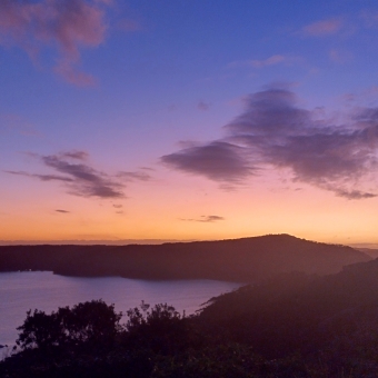 Flourescent Sunset, with the land below blackened into silhouette, with still water on the left which is ringed by land also which reflects the sky above, with the sky turning from navy blue at the top through magenta glowing orange near the horizon, with an occasional cloud appearing as a mauve silhouette against the sky