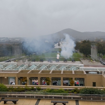 Blasted Cannon, New Parliament House, with the view from the top of New Parliament House over the Parliamentary Lawn where six miliary cannons are standing in a line anod pointing away, with the fourth cannon along in the middle of firing and an orange explosion is visible, with clouds of smoke visible rising in a general haze from the cannons, with the sky covered in grey rain clouds