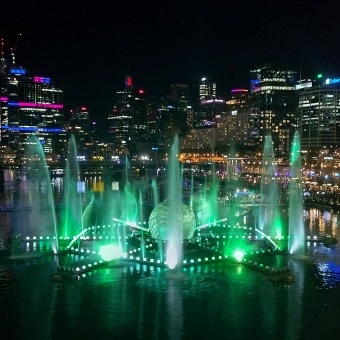 VIVID, Darling Harbour, Sydney, with the blackened sky above, with the high-rises of Barangaroo and Wynyard and Town Hall covering the horizon which stand out against the sky as their many luminous squares of windowed light mark their outlines, with a water feature about the size of a tennis court sitting in the middle of the waters of Darling Harbour which is spouting up water about five storeys in the air on all six sides of the water feature which is also lit up in a teal-green hue