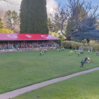 Cockington Green Football Club, with a stand full of fans undercover on the far side of the field on the left of the picture, with most of the football players on the right of the field and the image which includes a streaker, with normal size trees and a blue sky half covered in clouds visible in the distance