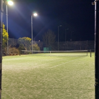 Court #1, Belconnen Tennis Courts, Belconnen, with a tennis court viewed from one corner and the net in the centre of the picture in the mid-foreground, with high fencing and floodlights around the edge, with the darkened sky above appearing navy blue because of the lightwash from the floodlights