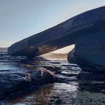 Avalon Headland, Avalon, with wet rocks in the bottom half of the image, with a sky beginning to glow at the beginning of a sunset which is about to start at the top of the image, with a large slanted rock leaning against another making a frame with a window thrugh which the light seems to glow more intensely