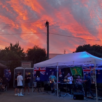 Band Out The Back, Adventure St at Moby Dick's Tavern, Kippax, with four musicians under a marquee lit up with LED lights, with people with beers in their hands watching and dancing, with red-glowing clouds from a sunset above