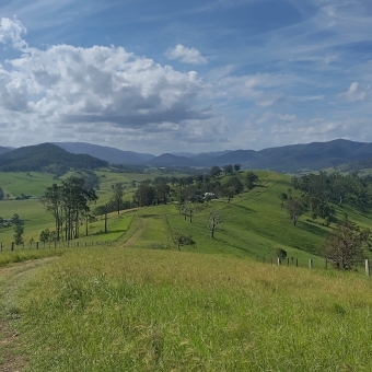 Kia Ora Lookout, Gloucester, with a rolling green hill sloping down into the valley occasionally dotted with green trees which all extends up into blue-hued mountains covering the horizon, with fluffly clouds near the horizon in an otherwise blue sky