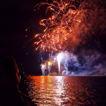 SkyFire, Regatta Point, Lake Burley Griffin, with two red bursting fireworks front and centre, with three violet fireworks shooting up from ground level, with these colours reflected in the seeming black water below, with a mobile phone taking a photo of the scene in the bottom left corner