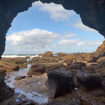 A Rocky Outlook, Caves Beach, with a cave opening framing the picture on all sides, with climbable boulders and rocks extending into the ocean, with patchy mashed potato clouds in the slightly visible sky