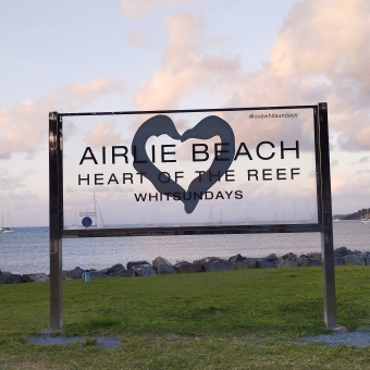 Airlie Beach Sign Over Sunset, with a transparent sign against the sunset which reads 'Airlie Beach, Heart of the reef, Whitsundays'