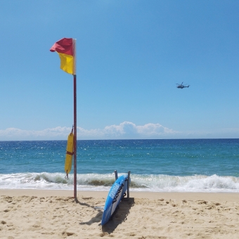 Chopper Protection, Coogee Beach, Coogee, with a black helicopter in the middle distance flying over the water, with a lifeguards' knee board and red-and-yellow flag on the sand