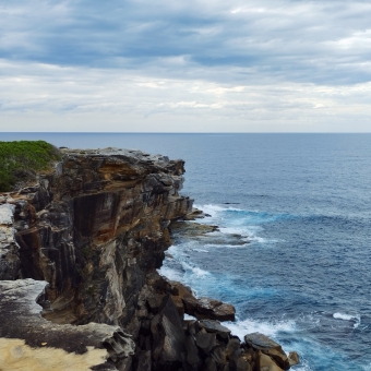 A Steep Dropoff, Headland Lookout, with cliffs adorned with occasional shrubbery on the left, with rippling ocean on the right crashing against each other