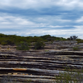Rocky Road Layer Cake, Boora Point, with the rocks rippling like leaning lasagne which has occasional shrubbery, with rolling clouds above which are a mix of blue and grey and yellow
