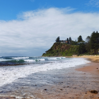 Bungan Head fron Newport Beach, Newport, with rippling and rough waves from left to right across an empty beach, with a cliffy headland in the distance, with a translucent blanket of cloud in the sky