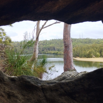 Masonic Frame, near Yeramba Lagoon, Picnic Point, with a wide hole in a rock cliff maing a frame for two tree trunks and a river and many trees lining the far bank