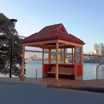 Peeking City, Henry Lawson Bus Stop, McMahons Point, with a bus stop frame coloured with cantaloupe and maroon in the foreground which has the Sydney Opera House peeking through its side and the Sydney Harbour Bridge from its left and CBD buildings from its right while all overlooking blue water reflecting a clear sky