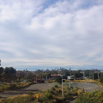 Tumultuous City Sky, IKEA, Tempe, with rippling grey clouds above green bushes and trees and the city skyline in the centre distance