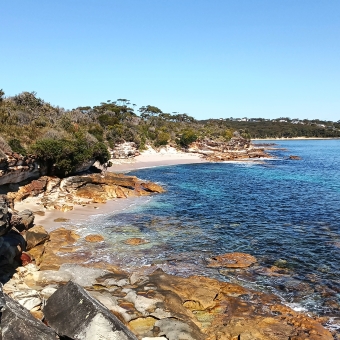 Hidden Swim, Little Jibbon Beach, Royal National Park, with a rocky foreshore leading up to a small beach in centre frame which continues into rocky foreshore into the distance, with green trees to the left and rippling blue water to the right
